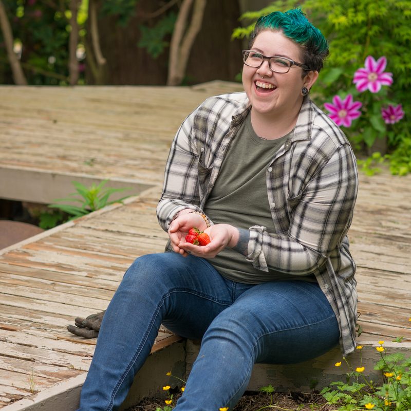 An agender person picks ripe red strawberries from a (moderately weed-laden) garden bed. Their preferred pronouns are they and them. They're wearing blue denim jeans, an olive green shirt, and a gray and white plaid flannel button-down shirt. They are also wearing glasses and ear plugs, and have bright blue-green dyed hair.