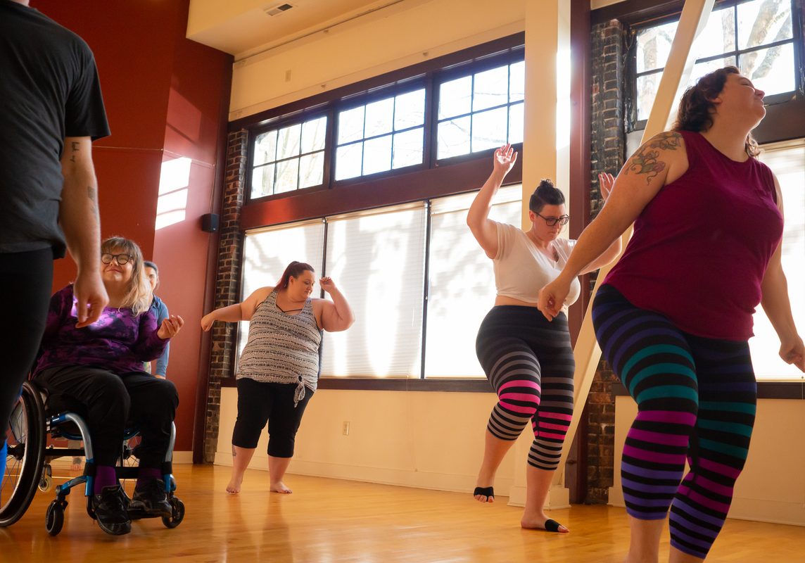 Students move, stretch, and dance in an inclusive dance class. The class includes people who are plus size or fat, people of color, and people with visible disabilities using mobility aids.